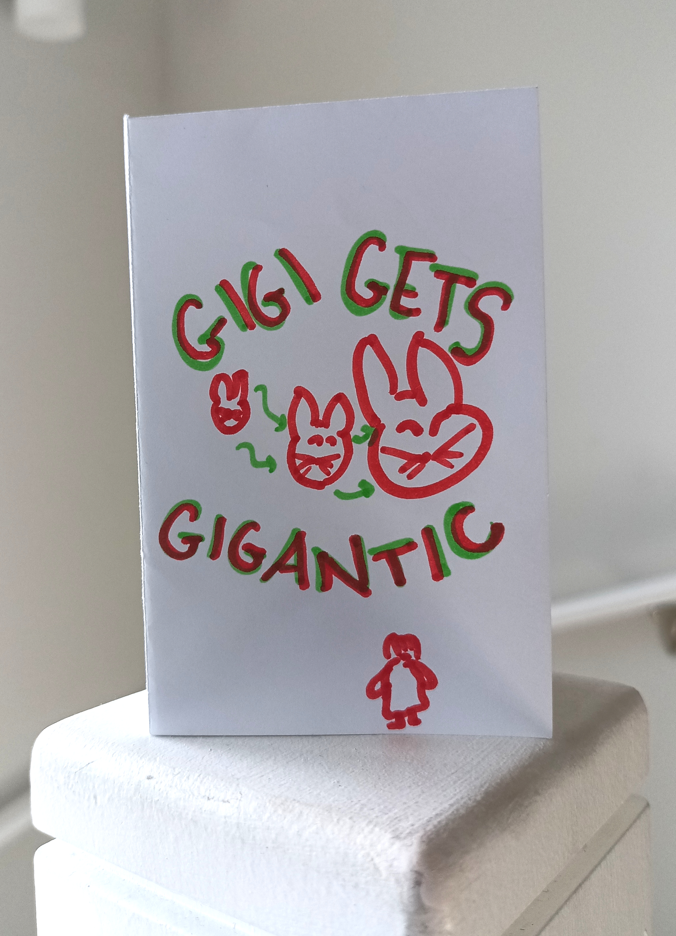 Photo of propped up zine drawn in red and green marker. Title Gigi gets gigantic. Gigi's cat face getting bigger and a figure in a dress at the bottom.